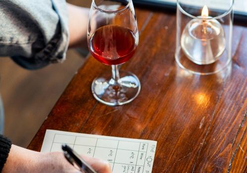 A glass of wine on a table and a hand taking notes on that table. 