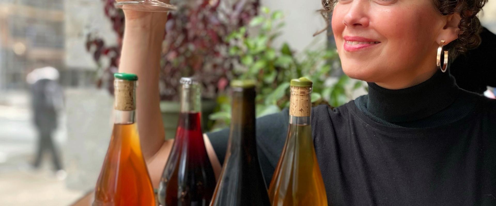 Heather Rankin holding a glass of wine, with 4 Lieux Communs wine bottles sitting on the ledge in front of her. 